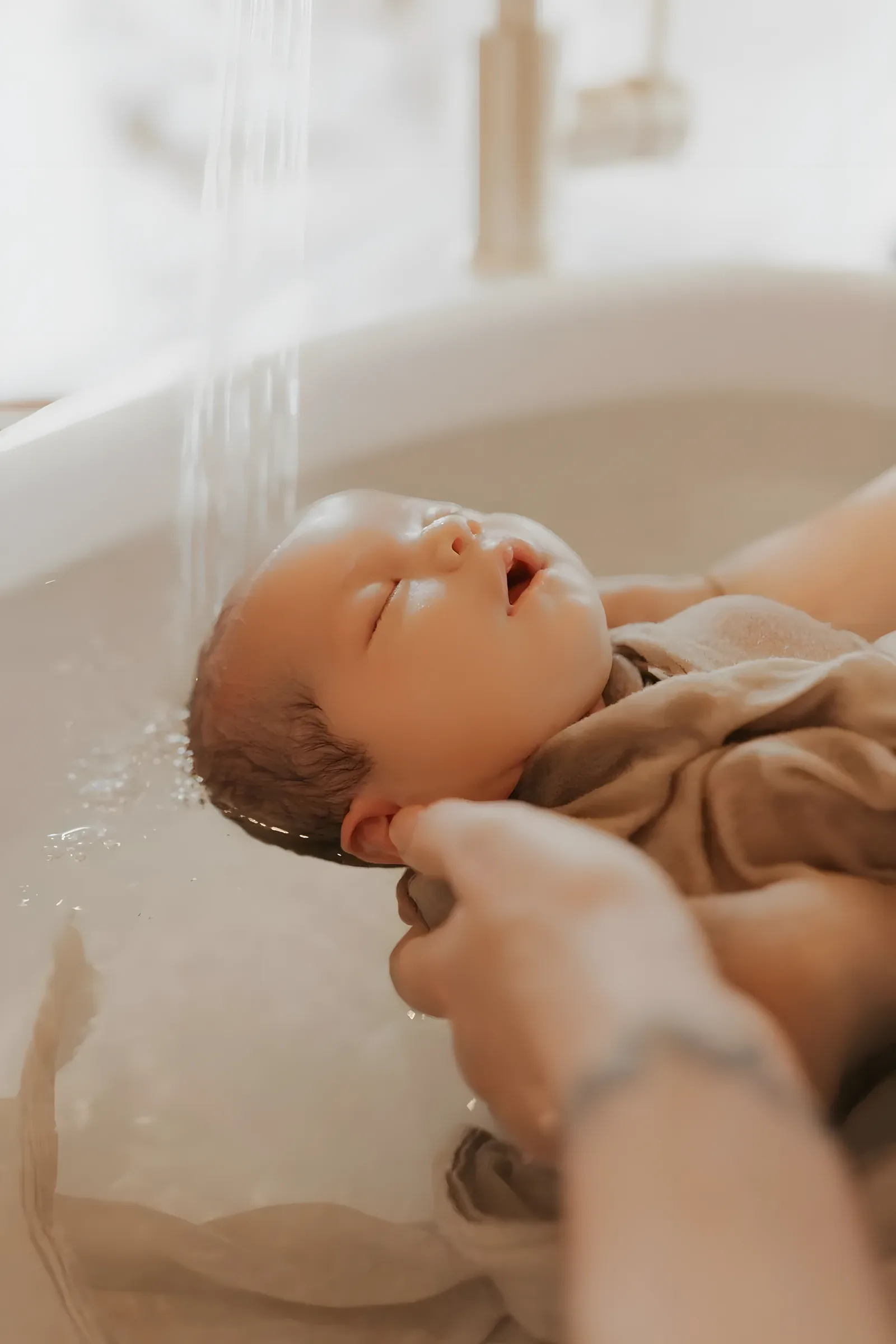 Nouveau-né enveloppé dans un lange, la tête soutenue par la praticienne sous un filet d’eau tiède, pendant le Bain d’Accueil du Nouveau-né®